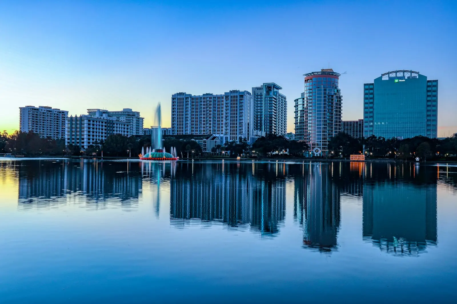 Alacakaranlıkta Orlando’da Lake Eola; gölün yüzeyinde yansıyan yüksek binalar ve ortadaki fıskiye.